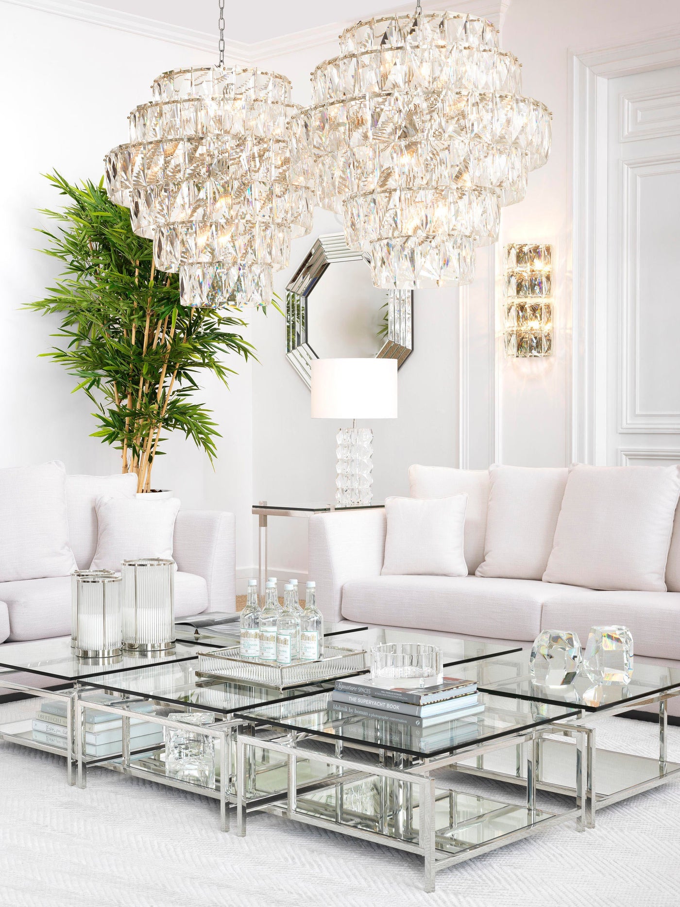 Eichholtz living room setting with elegant sofas, twin chandeliers, wall lights, mirror, glass and metal coffee tables and home accessories, styled to perfection.
